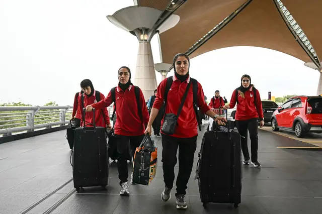 Altı futbolcu ellerinde bavullarıyla yürüyor. Üzerlerinde kırmızı üniforma ve siyah pantolon var.