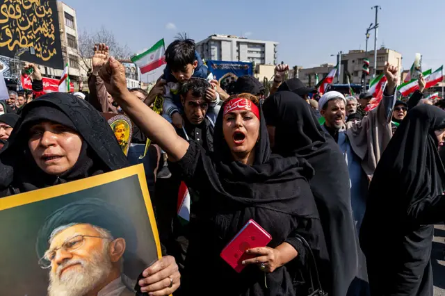 Önde Ayetullah Hamaney'in fotoğrafını taşıyan bir kadın. Arkada bir yumruğunu yukarı kaldırmış bağıran bir kadın var. Arkalarında yumruklarını kaldırmış insanlar görülüyor. İnsanların bir kısmı İran bayrağı taşıyor.