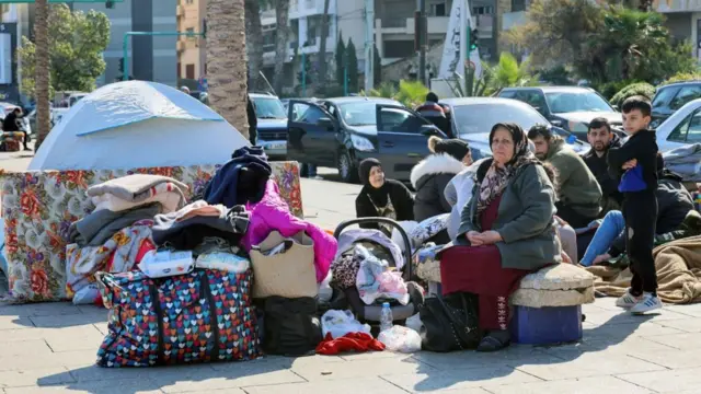 عائلات لبنانية من رجال ونساء وأطفال تجلس على الأمتعة في الشارع وخلفهم طابور من السيارات بعد النزوح من جنوب لبنان.