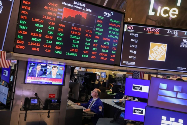 شاشات كبيرة ومتوسطة الحجم معلقة بمستويات مختلفة عليها مجموعة من الأرقام أو الأخبار، يظهر أحد الأشخاص في القاعة وكأنه يراقبها.