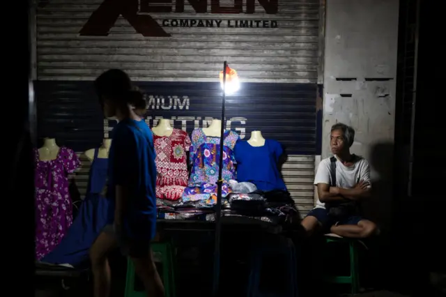 ၂၀၂၅ ခုနှစ်၊ ဖေဖော်ဝါရီလ ၁ ရက်မှာ မီးပျက်ချိန် ရန်ကုန်မြို့က အထည်ဆိုင်တစ်ဆိုင် မြင်ကွင်း၊ အမျိုးသမီး တစ်ဦး ဆိုင်ရှေ့က ဖြတ်သန်းသွားတာတွေ့ရပြီး ဆိုင်ရှင်လို့ ယူဆရတဲ့အမျိုးသားတစ်ဦး လက်ပိုက်ပိုင်း အဝေးကို ငေးကာ ထိုင်ခုံပေါ် ထိုင်နေ