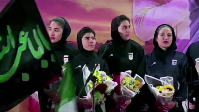 kadın futbolcular kürsüdeler, ellerinde de çiçekler var.