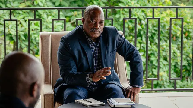 Joseph Kabila yicaye arimo kuvuga yambaye ikositimu n'ishati n'ipantalo y'ikoboyi, akoze ikimenyetso n'ikiganza. Imbere ye ku meza hari amakayi n'ikaramu. Imbere ye haraboneka umutwe w'umuntu wicaye umukurikiye. Ifoto yafotorewe aho aba i Goma, hagenzurwa n'inyeshyamba za M23, mu burasirazuba bwa DRC, ku itariki ya 30 Gicurasi (5) mu mwaka wa 2025.