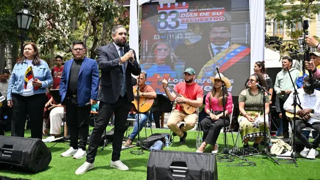 Nicolás Maduro Guerra, vistiendo traje y corbata, y zapatillas blancas, en un pequeño estrado hablando por un micrófono. Detrás hay músicos sentados.