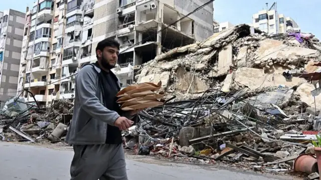 شاب يسير في منطقة في بيروت، حيث تظهر مبانٍ سويت بالأرض على جانب الطريق