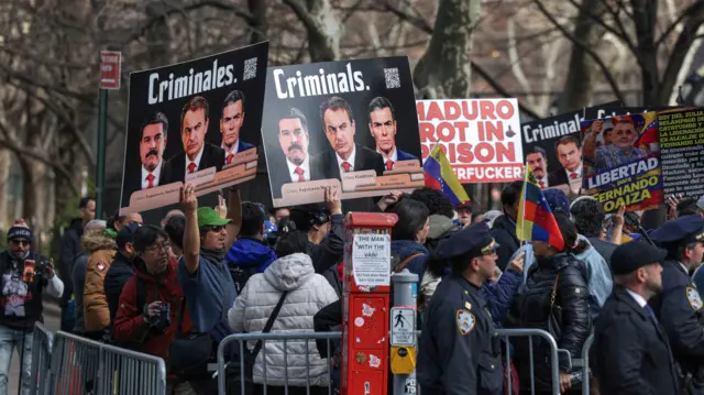 Policías detrás de una valla y, en el fondo, manifestantes con carteles de "criminales" y las fotos de Maduro, Zapatero y Pedro Sánchez.