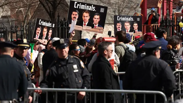 Policías detrás de una valla y, en el fondo, manifestantes con carteles de "criminales" y las fotos de Maduro, Zapatero y Pedro Sánchez.