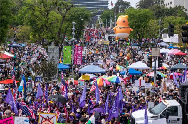 حشد كبير من المتظاهرين يحملون أعلاماً بألوان مختلفة إلى جانب العلم الأمريكي، وشعارات مناهضة لترامب. ووسط الحشود تظهر مركبة بيضاء اللون.