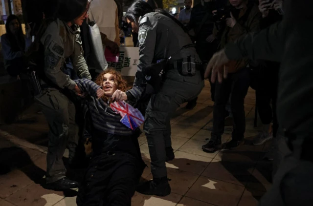 شرطيتان تعتقلان محتجة شعرها يميل إلى النحاسي وترتدي بنطالاً أسود وقميصاً يبدو رمادياً مخططاً باللون الكحلي. الصورة التقطت ليلاً.