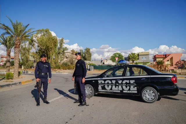 صوة أرشيفية لقوات أمن عراقية تقوم بحراسة طريق بشمال مدينة الموصل العراقية، ويقف فردان تابعان للشرطة العراقية مدججان بالسلاح ويرتديان زياً شرطياً أسود، وخلفهما سيارة سوداء كتب عليها كلمة "شرطة" باللغة الإنجليزية.