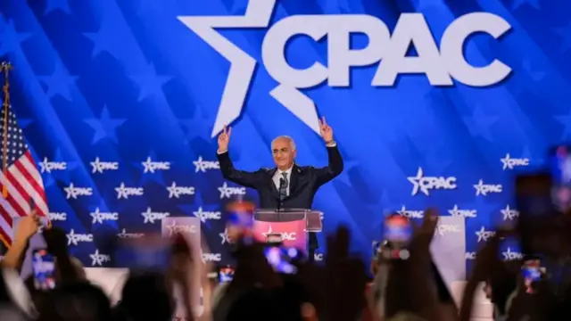 رضا بهلوي يرتدي بدلة رسمية يبدو أن لونها أسود مع قميص أبيض وربطة عنق، خلفه العلم الأمريكي، ويحيي أنصاره في مؤتمر العمل السياسي المحافظ في ولاية تكساس الأمريكية