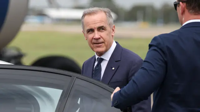 El primer ministro canadiense, Mark Carney, vistiendo un traje azul y una camisa clara mira a un hombre que está de espaldas abriéndole la puerta de un vehículo para que él entre.