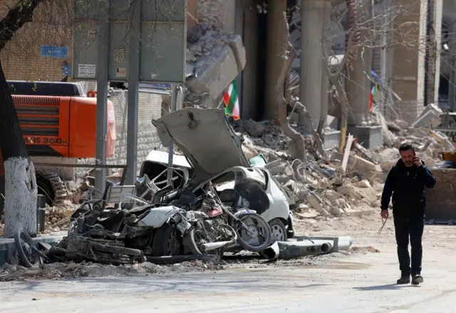 житель Тегерана на фоне разрушенного здания и разбитого автомобиля