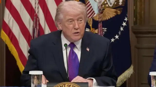 ترامب خلفه العلم الأمريكي وأمامه مايكروفون ويتحدث. يرتدي بدلة رسمية تبدو سوداء اللون مع ربطة عنق بنفسجية وقميص أبيض مع دبوس صغير للعلم الأمريكي وأمامه كأس ماء.