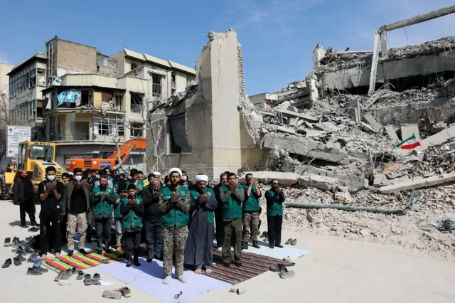 жители Тегерана молятся на фоне разрушенного в результате авиаудара здания