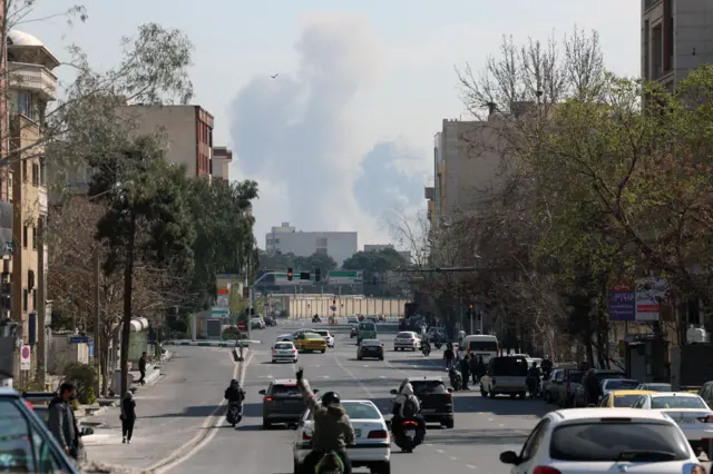 движение в городе, на заднем плане видны столбы дыма на месте удара