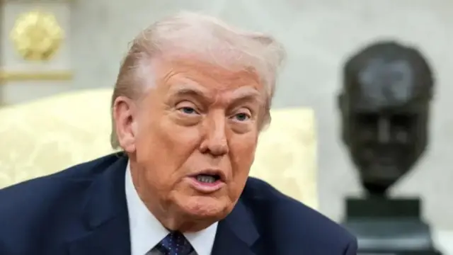 الرئيس الأمريكي دونالد ترامب يرتدي بدلة رسمية كحلية اللون ويبدو أنه يتحدث ويجلس على مقعد.