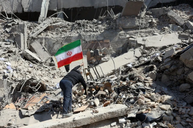 إيراني يضع العلم الوطني بين أنقاض مبنى مركز شرطة متضرر في وسط طهران، يبدو رجلاً كبيراً في السن يرتدي بنطالاً كحلي اللون وقميصاً أسود، يظهر منحنياً من الخلف. الصورة التقطت نهاراً.