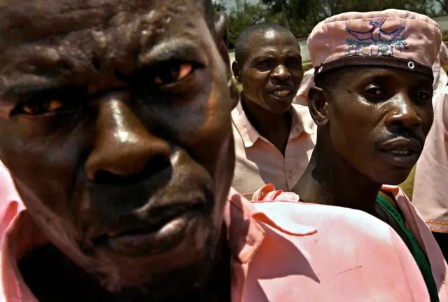 Imfungwa eshatu z'abagabo zo mu Rwanda, zambaye imyenda y'iroza iziranga, umwe muri bo yambaye ingofero y'iroza.