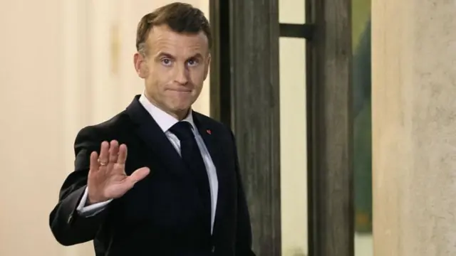 Emmanuel Macron de terno escuro acena em direção à câmera com testa franzida e lábios cerrados