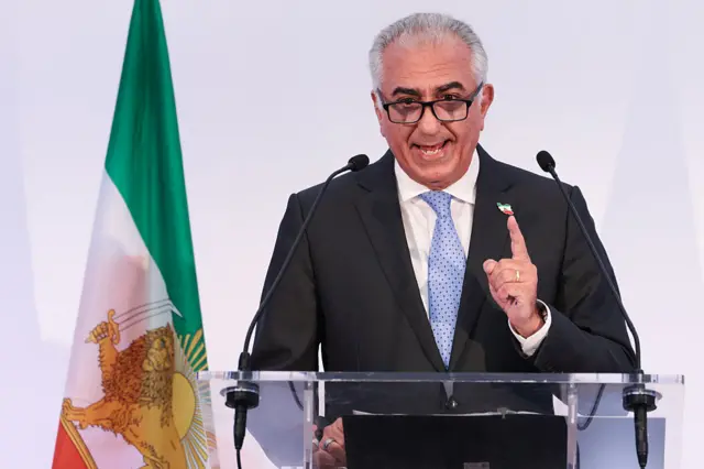 رضا پهلوی پشت تریبون در حال ایراد سخنرانی است. پرچم ایران با نشان شیر و خورشید در کنار او قرار دارد.