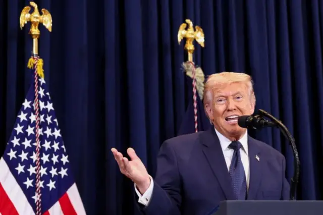 Trump kürsüde sağ elini kaldırmış konuşurken arkasında bir ABD bayrağı görülüyor