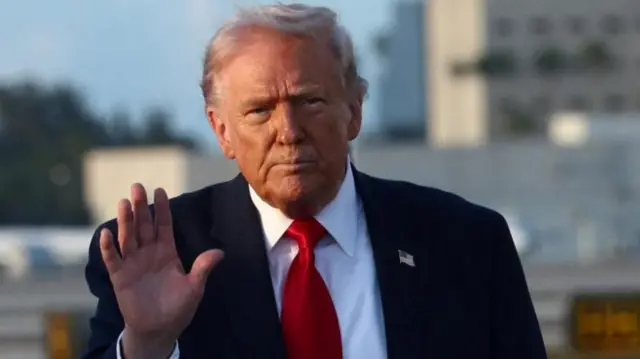 Trump üzerinde takım elbiseyle elini kaldırmış kameraya doğru bakıyor