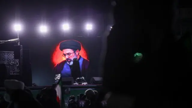 Mücteba Hamaney'in duvara asılmış bir fotoğrafı uzaktan çekilmiş, önünde insanlar görülüyor