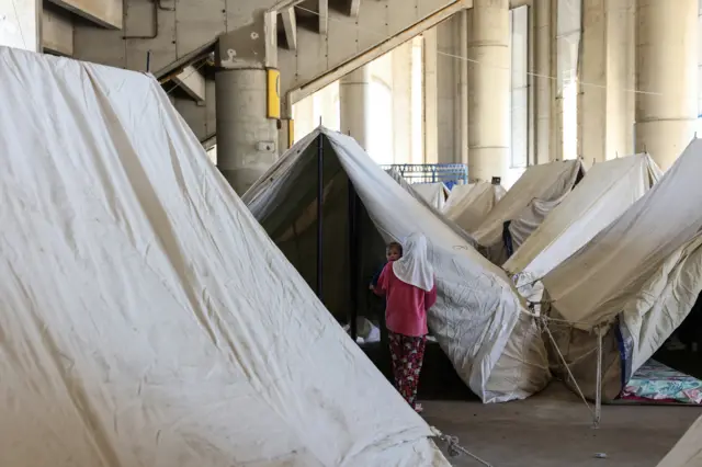 امرأة نازحة تحمل طفلها أثناء سيرها بين الخيام داخل استاد مدينة كميل شمعون الرياضية، الذي حُوِّل إلى مركز استقبال وإيواء للنازحين، في بيروت بتاريخ 10 مارس/آذار 2026.