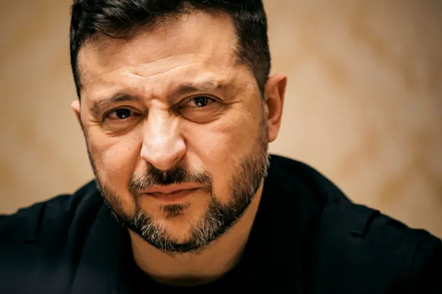 زيلينكسي يرتدي قميصاً أسود اللون، بشرته فاتحة وشعره غامق، ولحيته سوداء ورمادية.