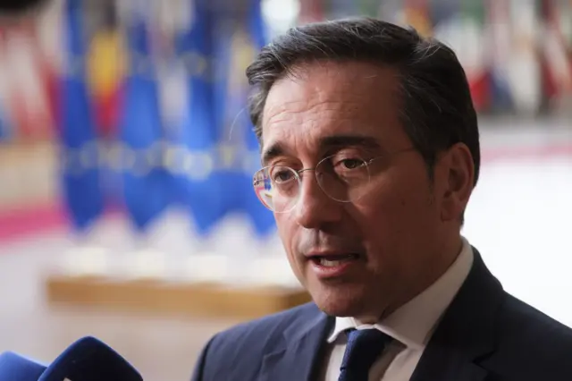 بوينو يرتدي بدلة رسمية كحلية اللون وقميصاً أبيض وربطة عنق كحلية، ونظارة طبية. شعره بلونين أسود ورمادي وبشرته فاتحة، ويبدو أنه وراءه أعلام.