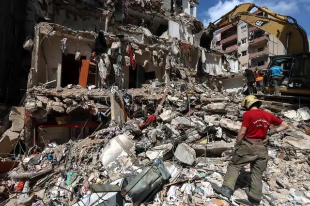 লেবাননের বৈরুতের উপকণ্ঠে ১০শে এপ্রিল ইসরায়েলি বিমান হামলায় আংশিকভাবে ধ্বংস হওয়া একটি ভবনের ধ্বংসস্তূপ সরাতে বুলডোজার নিয়ে কাজ করছেন উদ্ধারকর্মীরা