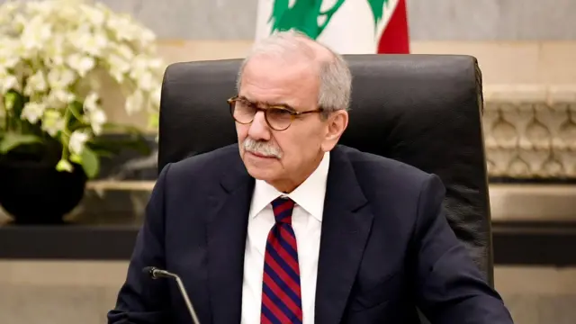 نواف سلام نشسته روی صندلی سیاه با کت سرمه‌ای و کراوات قرمز