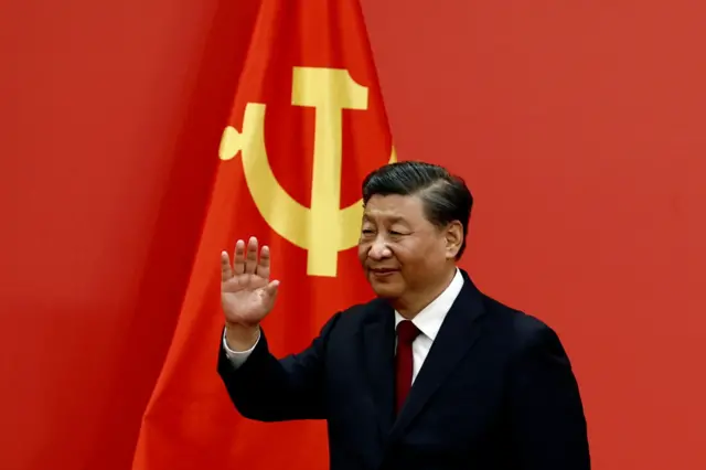 Ifoto ya Prezida w'ubushinwa Xi Jinping, azamuye ikiganza. Yambaye ikote ry'umukara.