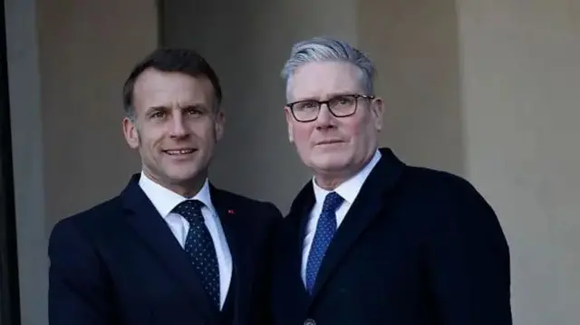 Perezida w'Ubufaransa Emmanuel Macron ahagararanye na Minisitiri w'intebe w'Ubwongereza Keir Starmer. Bambaye ikositimu zijimye hamwe na karuvati.