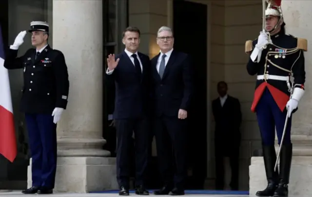 ستارمر يقف إلى جانب ماكرون وأمامهما حارسان. كلا المسؤولين يرتديان لباسا رسميا ويلوح ماكرون بيده بينما يحتضن ستارمر ماكرون بيده اليمنى.