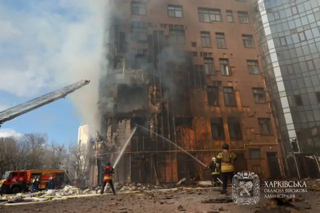 Многоэтажное здание, перед ним пожарные тушат возгорание.
