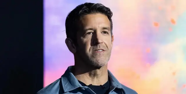 John Ternus arimo kureba muri 'camera' ahagana ku ruhande. Yambaye umwenda w'ubururu, imbere harimo undi w'umukara.