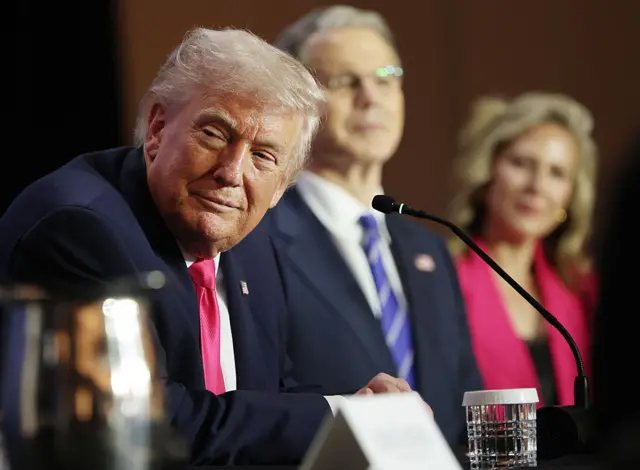 ترامب يرتدي بدلة رسمية كحلية اللون وربطة عنق تبدو بلون زهري غامق وقميص أبيض وأمامه مايكروفون وكوب ماء، وبجانبه رجل وامرأة ملامح وجهيهما غير واضحة.