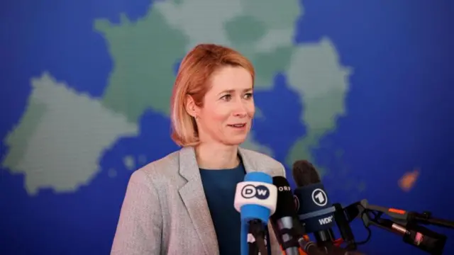 كاجا كالاس، بشعرها البني القصير، ترتدي بدلة رمادية، تقف أمام خريطة لأوروبا، وأمامها ميكروفونات.