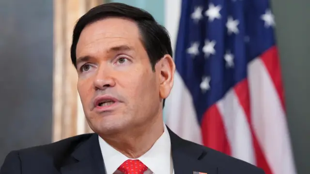 مارکو روبیو با کت سیاه و کراوات قرمز جلوی پرچم آمریکا