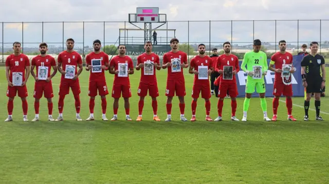 تیم ملی فوتبال ایران با لباس ورزشی روی زمین چمن و عکس دانش‌آموزان کشته‌شده در مدرسه میناب در دست