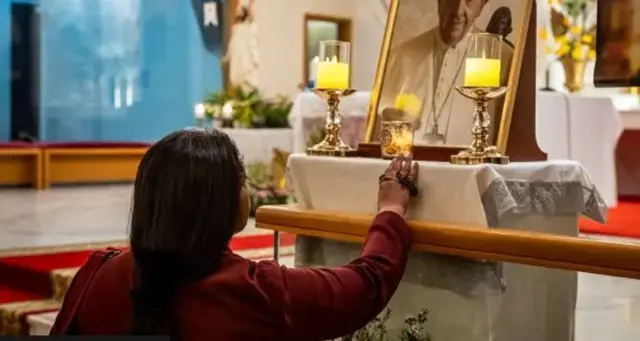 Ifoto yafatiwe inyuma y'umuntu uri mu Kiliziya apfukamye imbere y'ifoto ya Papa.