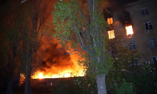 Пожар на заднем плане, на переднем плане дерево с зеленой листвой и многоэтажный дом, последние два этажа его тоже горят.