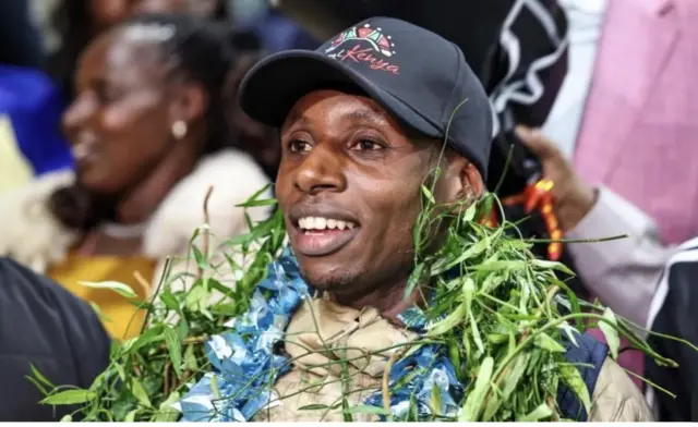 Sawe mu ifoto aseka akikijwe n’abaje kumwakira yambitswe mu ijosi ibyatsi mu kumwishimira, yambaye n'ingofero y’umukara.