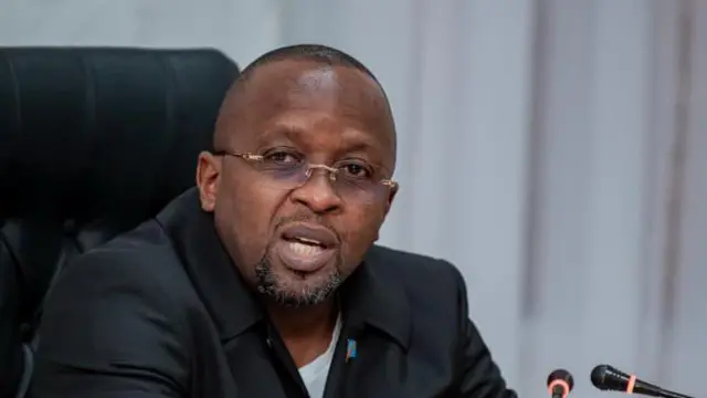 Minisitiri w'ubutegetsi bw'igihugu wa DRC Jacquemain Shabani arimo kuvugira mu ndangururamajwi ebyiri. Yicaye areba muri 'camera'.