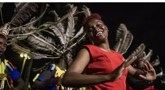 Ifoto igaragaza umwe mu babyinnyi wambaye akenda gatukura hejuru inyuma ye hari abandi babyinnyi bambaye gakondo baserukana mu kwizihiza ibirori