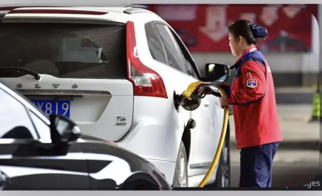 Ifoto y’imodoka y’umweru iri kunywa igitoro kuri 'station' n’umugore wambaye umupira w’umutuku uri guha iyo modoka igitoro.