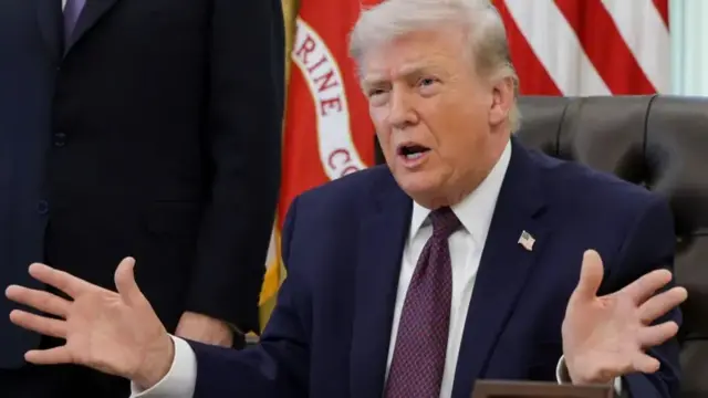 الرئيس الأمريكي دونالد ترامب يرتدي بدلة رسمية كحلية اللون وقميصاً أبيض وربطة عنق بنقط توتية وكحلية، يحرك يديه إلى الأمام متحدثاً ويجلس على كرسي أسود اللون.