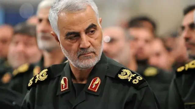 سليماني مرتدياً بدلة عسكرية لونها زيتي، بشرته فاتحة لكنها تميل إلى اللون القمحي ولحيته قصيرة لونها أبيض وشعره لونه أبيض.